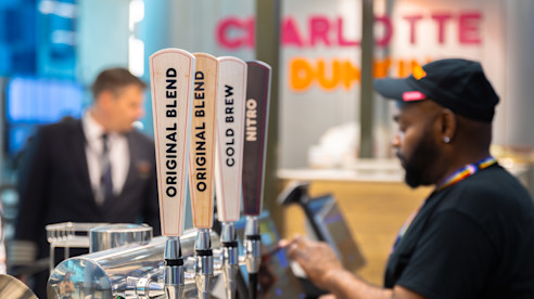 Mostrador de cafetería con grifos etiquetados Original Blend, Cold Brew y Nitro, mientras un barista atiende a un cliente.