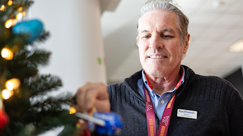 Hombre decora un árbol navideño en interior, colocando un adorno entre luces y ramas.