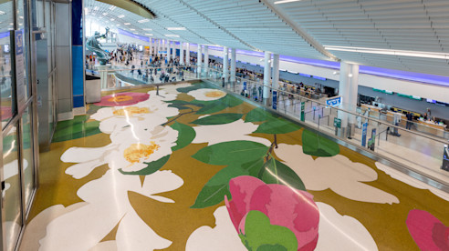 Vista amplia de un terminal de aeropuerto con un gran diseño floral en el piso con flores blancas y rosas, y viajeros avanzando por las filas de seguridad.