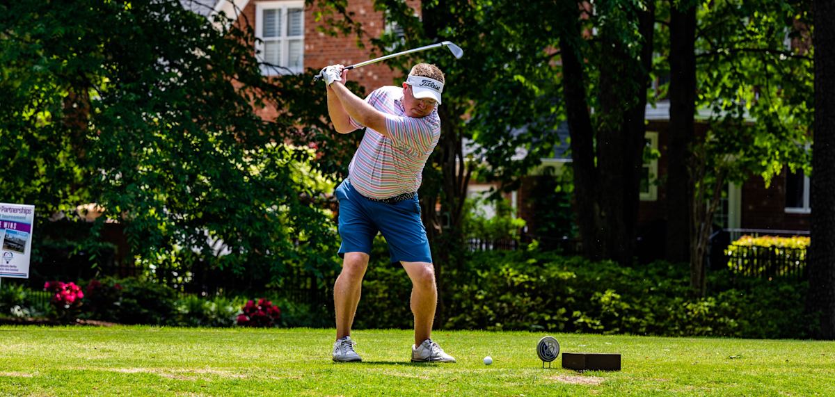 A man prepares to hit a golf ball on a green golf course. He is wearing a striped shirt, blue shorts, and a white visor while swinging a golf club. Trees, flowers, and brick houses are visible in the background on a sunny day.