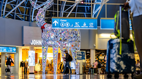 Gran escultura de reno iluminado en la terminal del aeropuerto mientras pasajeros caminan hacia tiendas y puertas.