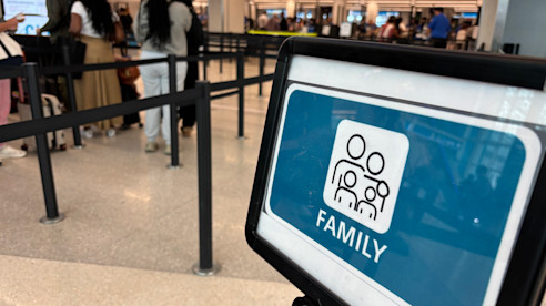 Close-up of a “Family” sign at airport security lane with families waiting in line in the background.