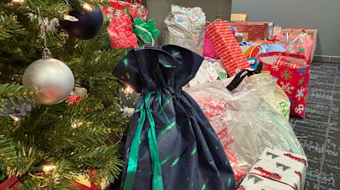 Regalos envueltos y bolsas de donaciones están apilados junto a un árbol de Navidad decorado en interior.