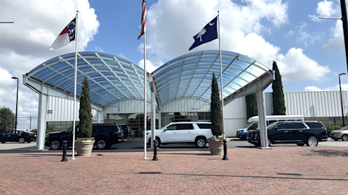Glass-canopy airport entrance with flags and parked vehicles in front of a brick-paved driveway.