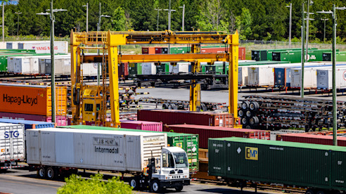Patio de carga intermodal con contenedores, camiones y una gran grúa amarilla moviendo mercancía cerca de las vías del tren.