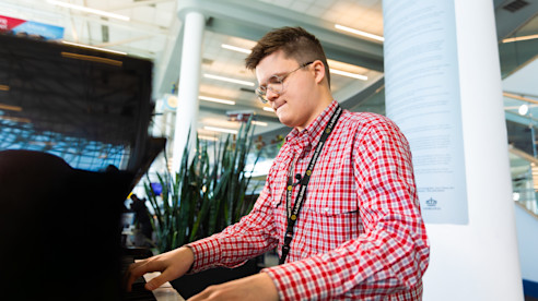 Man playing a piano inside an airport terminal with seating and signage visible in the background.