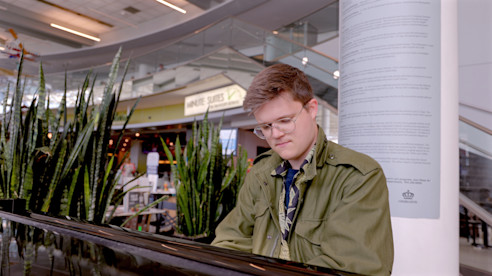 Josh King playing a piano inside an airport terminal with plants and seating nearby.