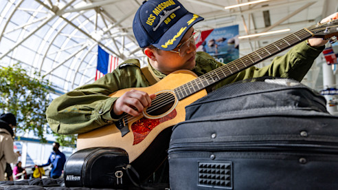 Josh King playing an acoustic guitar inside an airport terminal with luggage nearby.