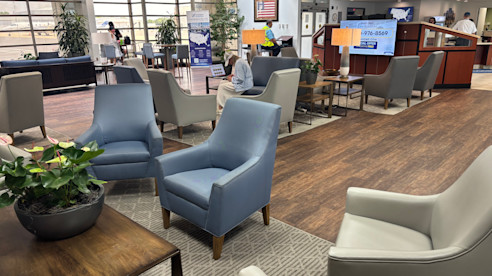 Spacious airport lounge with modern seating, natural light, potted plants, and passengers relaxing.