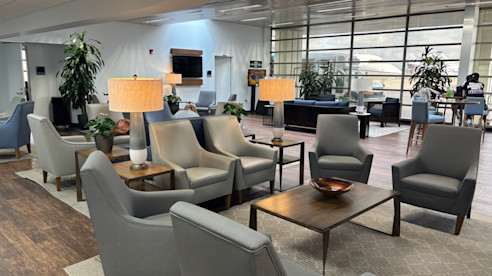 Modern airport lounge with gray armchairs, coffee tables, and warm lighting in a spacious, open setting.
