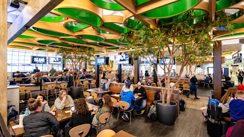 Ajetreada cervecería al aire libre en el aeropuerto, con viajeros comiendo en mesas de madera, rodeados de árboles interiores y un moderno diseño de techo.