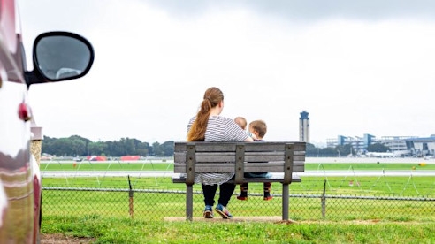 Mujer y niño sentados en una banca observan los aviones cerca de la pista del Aeropuerto CLT, con la torre de control visible a lo lejos.