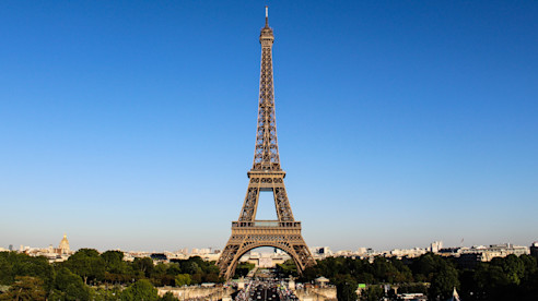 View of the Eiffel Tower in Paris rising above the city with trees and buildings in the foreground.
