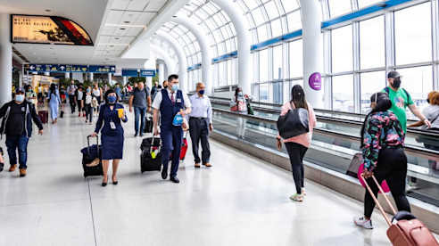 Viajeros caminan por una terminal luminosa del aeropuerto con grandes ventanales; algunos usan la pasarela móvil mientras otros llevan equipaje y usan mascarillas.