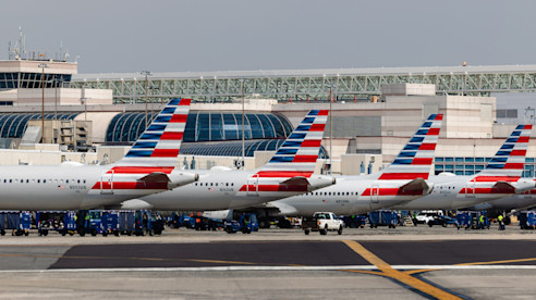 A lineup of American Airlines planes at the airport, with the control tower and terminal buildings in the background, highlighting the busy hub operations.