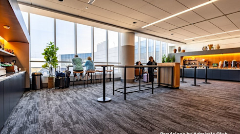 Airport lounge with food service area, seating by tall windows, and passengers relaxing with views outside.
