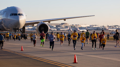 Participants running in an airport runway 5K event, passing by a large parked airplane with terminals in the background.