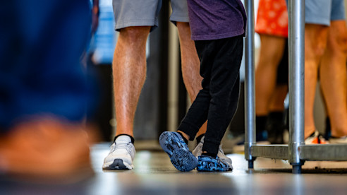 Close-up of child standing beside an adult in airport security line, focused on feet and lower legs.
