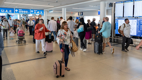 Busy airport terminal with travelers walking through the concourse and flight information displays overhead.