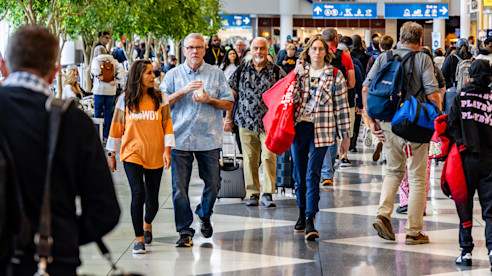 Crowded airport concourse with travelers walking past indoor greenery and overhead directional signs.