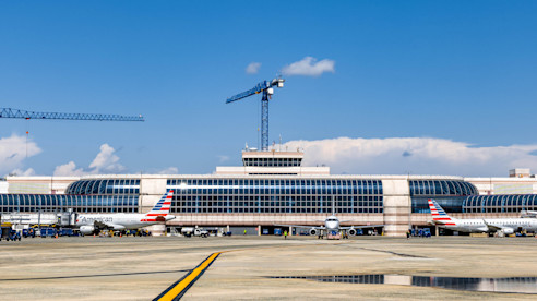 Wide view of an airport terminal with multiple aircraft at gates and construction cranes visible in the background.
