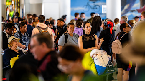 Crowded airport terminal with travelers standing closely together, some holding bags and personal items.