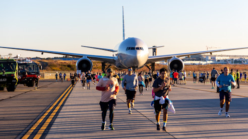 Corredores participan en una carrera sobre la pista del aeropuerto con un avión grande al fondo.