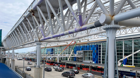 Airport terminal exterior with elevated pedestrian walkway, steel supports, and traffic moving below.
