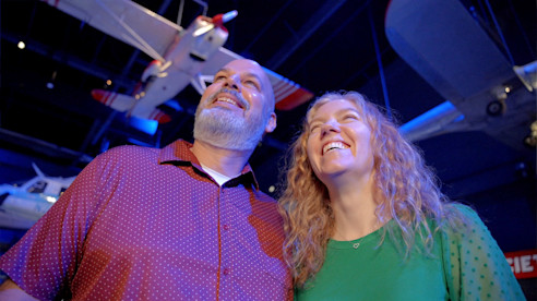 Two adults smiling and looking up at aircraft exhibits inside an aviation museum.