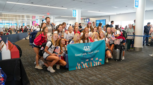 Team USA Women's Field Hockey players gather at the airport holding a "Good Luck, Team USA!" banner.