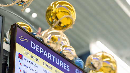 Pantalla de salidas con información de vuelos y logotipos de aerolíneas, con esferas doradas decorativas arriba.