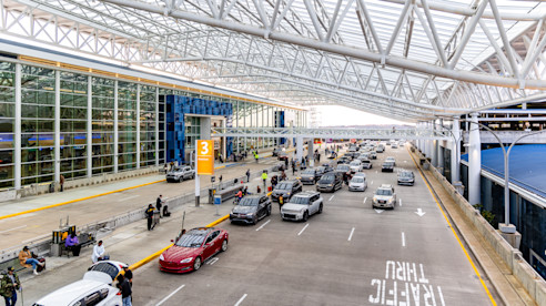 Airport terminal curbside with multiple lanes of vehicles, passengers loading luggage, and a covered drop-off area.
