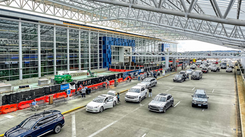 Busy airport drop-off area with cars, passengers, and ongoing terminal construction under a large glass and steel canopy.