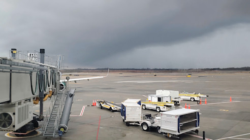 Zona de embarque del aeropuerto y pista bajo nubes de tormenta oscuras, con vehículos de servicio en tierra estacionados cerca del puente de embarque.