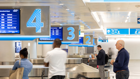 Pasajeros esperan en la zona de reclamo de equipaje en un terminal de aeropuerto, junto a carruseles marcados 2, 3 y 4 con pantallas de información de vuelos arriba.