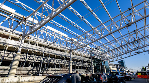 Steel canopy structure over an airport roadway with vehicles below and terminal buildings in the background.