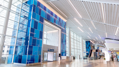 Amplio terminal de aeropuerto con techos altos blancos, grandes ventanales de vidrio y una pared azul llamativa cerca de una entrada.