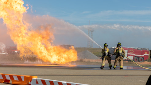 Bomberos realizan un ejercicio de entrenamiento con fuego en una pista del aeropuerto, usando equipo de seguridad.