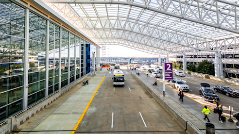 Carretera en el nivel superior del aeropuerto con automóviles, autobuses y pasajeros bajo una gran marquesina de vidrio y acero cerca de la entrada de la terminal.