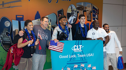 USA Taekwondo Team celebrates at the airport, holding a "Good Luck, Team USA!" banner with American flags.