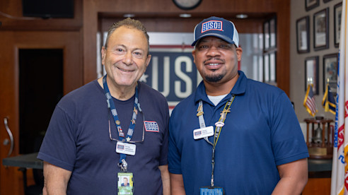 Two USO staff members smile in front of the welcome desk, wearing uniforms
