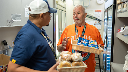 Two USO volunteers talk while holding trays of snacks and pastries in a supply room stocked with food and drinks.