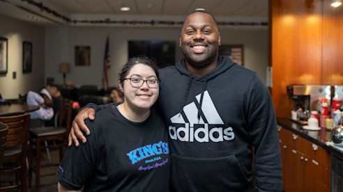Two people smiling and posing together inside a lounge or break area with a U.S. flag in the background.