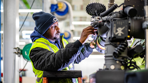 Trabajador de mantenimiento del aeropuerto con chaleco de seguridad y gorro repara equipo dentro de un taller utilizando herramientas especializadas.