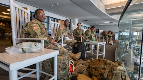 Uniformed service members sit on high stools with backpacks beside them in an airport terminal seating area.