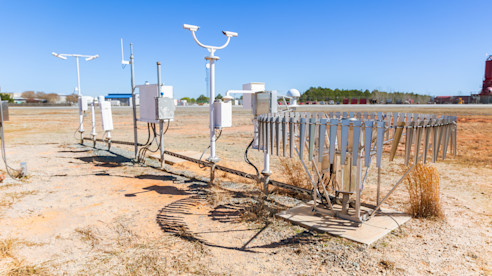 Equipo de monitoreo meteorológico con varios sensores e instrumentos instalados en un terreno cerca de la pista del aeropuerto bajo un cielo azul despejado.