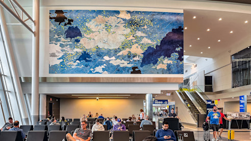 Airport seating area with passengers waiting, featuring a large abstract mural titled "Where Earth Meets the Sky" by Val Britton, depicting a dynamic blue and white cloudscape.