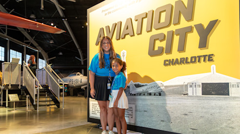 Dos niñas con camisetas azules posan sonrientes frente al cartel de “Aviation City Charlotte”