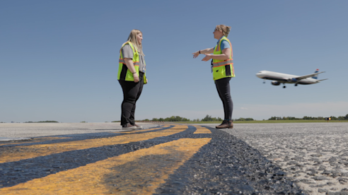 Dos trabajadoras del aeropuerto con chalecos de seguridad conversan en la pista mientras un avión despega al fondo.